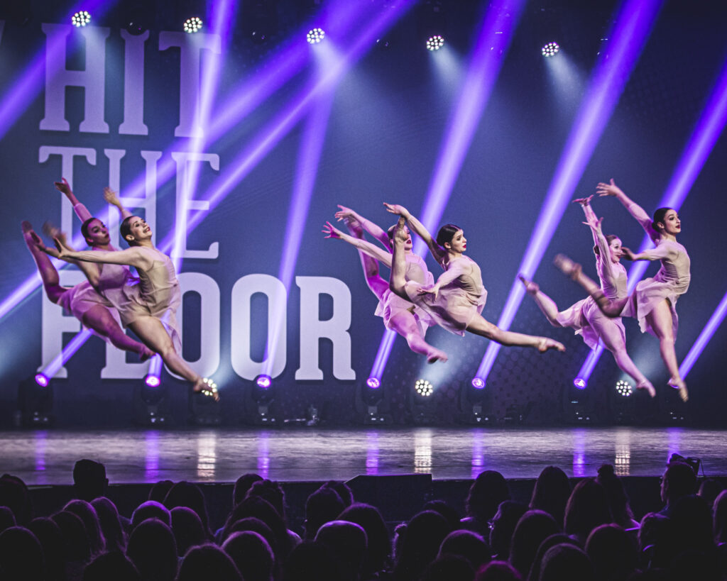 Des danseurs effectuent un grand écart en sautant sur une scène, où l'on peut voir Hit The Floor écrit derrière
